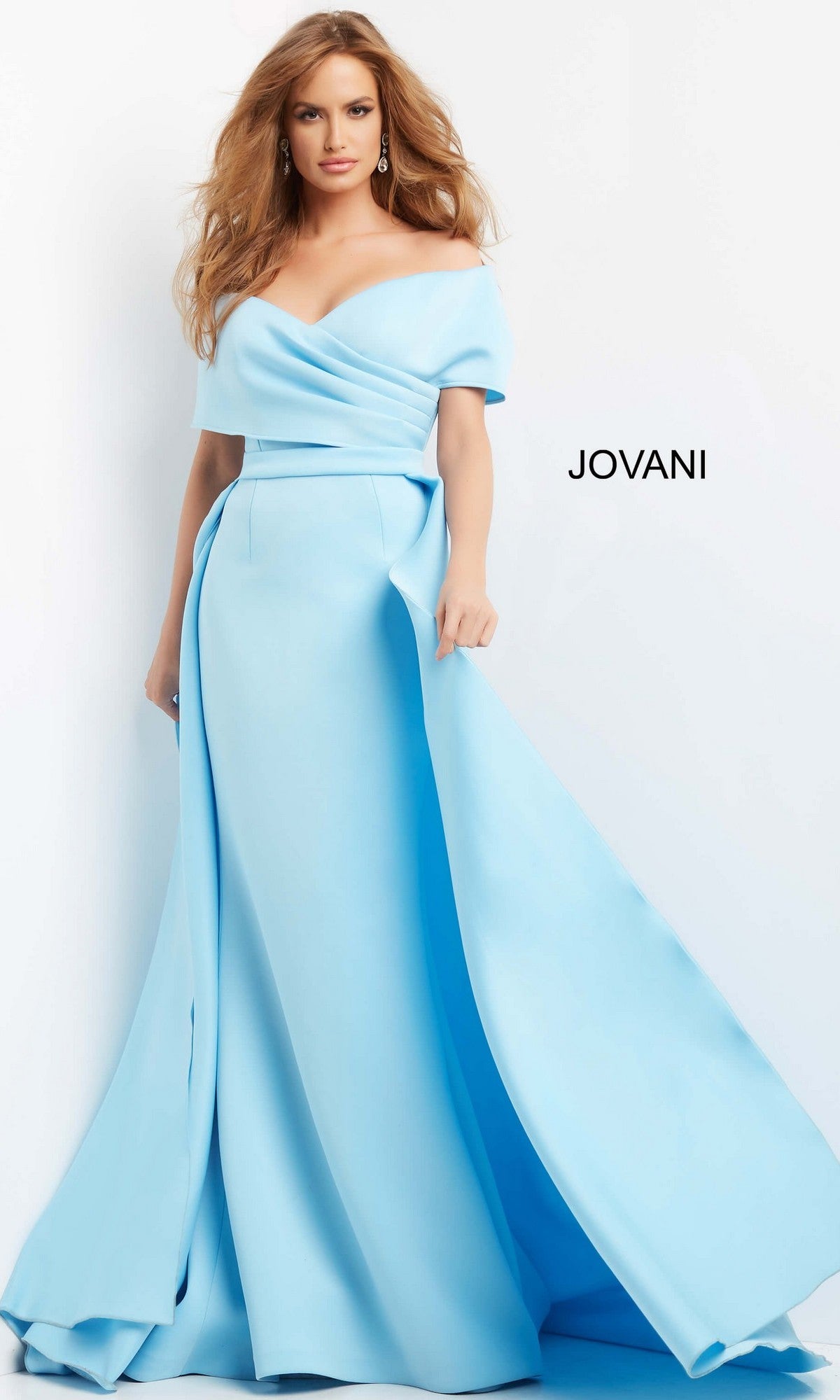 A woman wearing the Jovani 07443 Long Formal Dress—a light blue, off-the-shoulder, floor-length gown with a full skirt—stands beside a wall with “JOVANI” displayed on it.